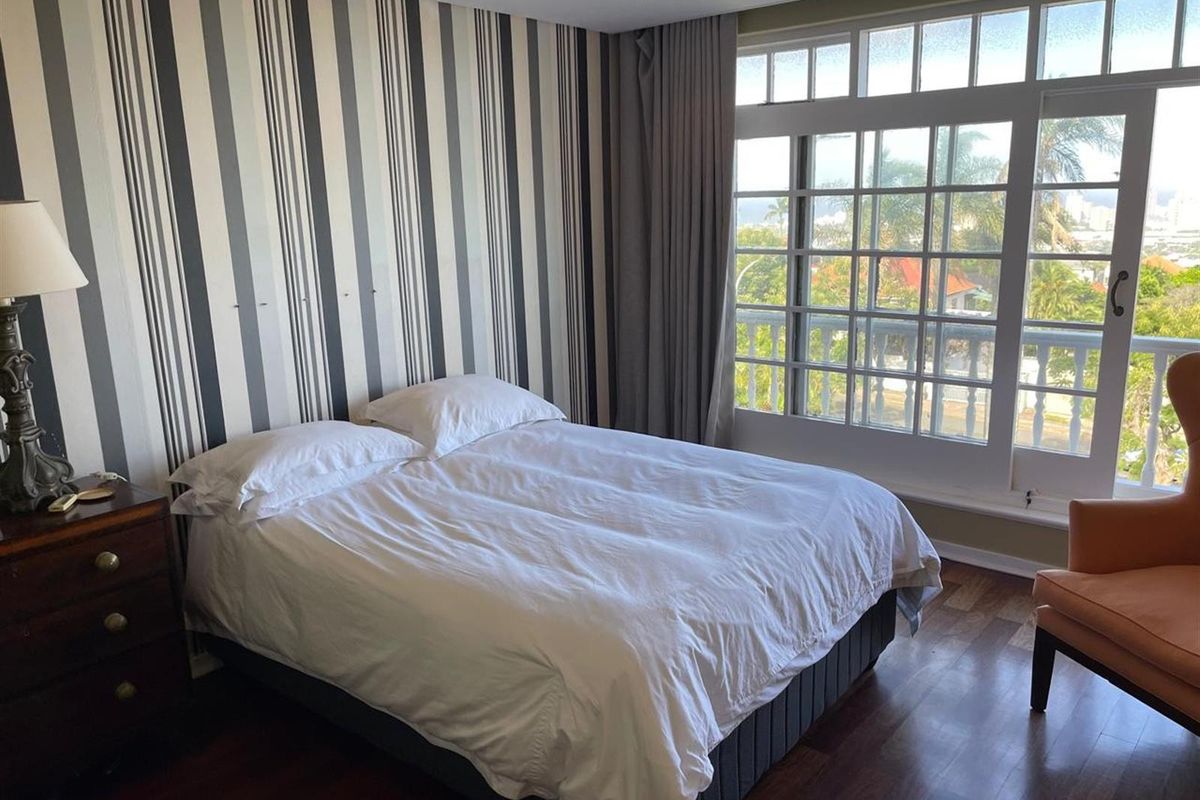 A well-lit bedroom featuring dark wood flooring, a large multi-pane window with balcony access, and a feature wall with vertical striped wallpaper.