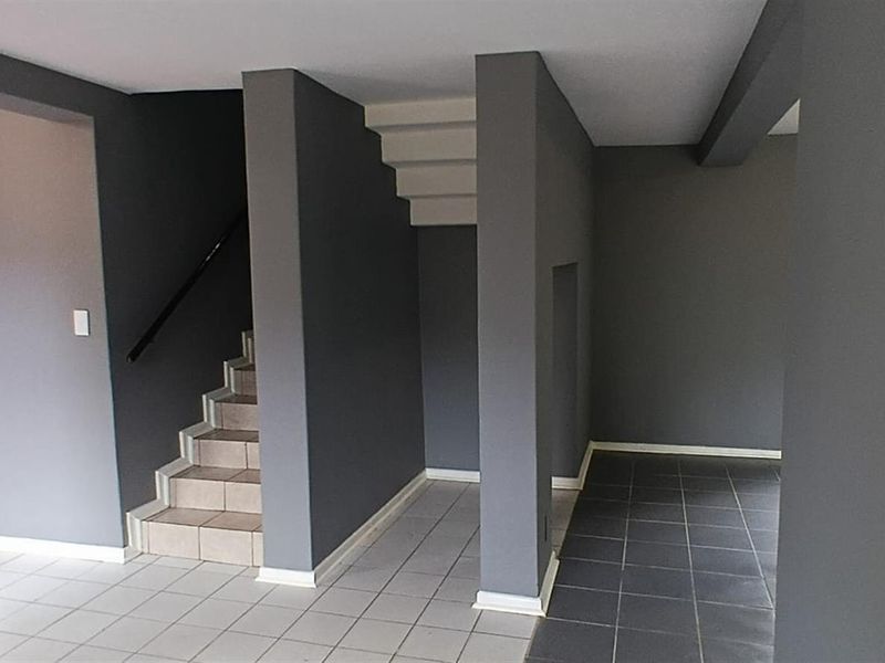 An open-plan entry area featuring a staircase, neutral grey wall finishes, and tiled flooring, leading into an adjacent kitchen space.