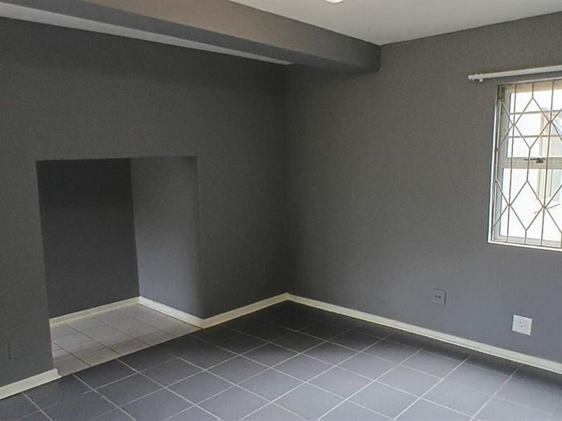 A clean, empty living room featuring dark grey tiled flooring, neutral walls, and a window fitted with security bars.