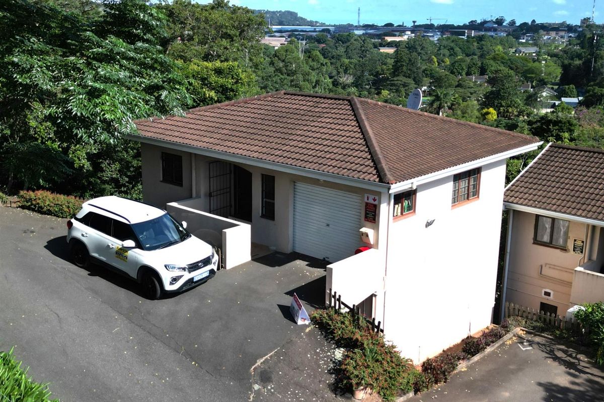Aerial view of a townhouse featuring a tiled roof, single garage, and paved driveway, set within a lush, elevated residential environment.