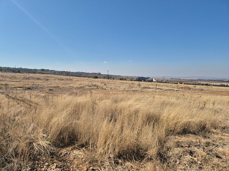 A wide-angle view of a vacant residential plot featuring dry grass and distant landscape views under a clear blue sky.