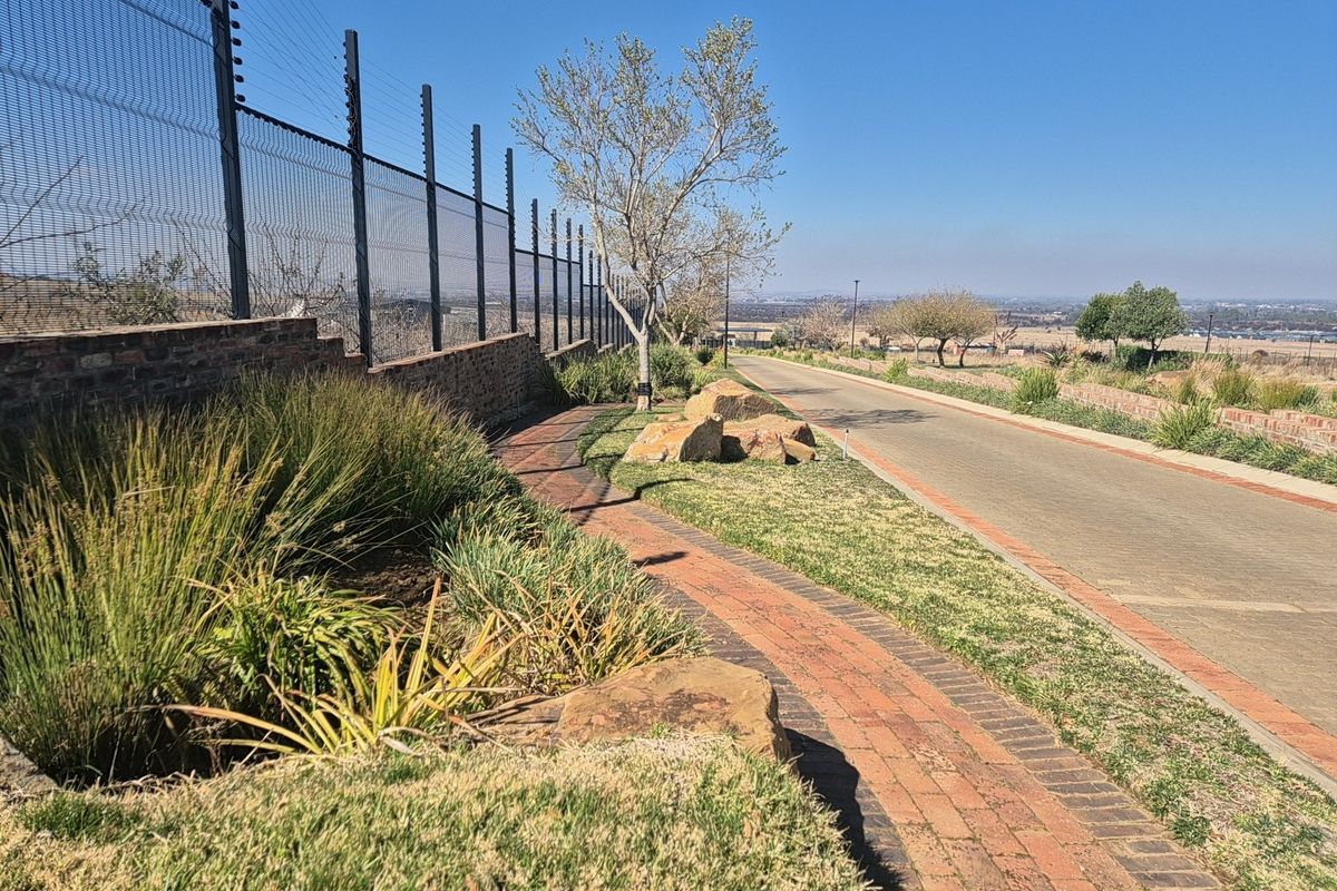 A well-maintained residential street view featuring a paved walkway, brick retaining wall, electric fencing, and landscaped garden areas.
