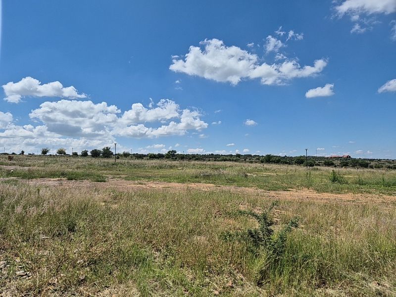A wide-angle view of a vacant residential plot featuring natural grassland and perimeter fencing under a clear blue sky.