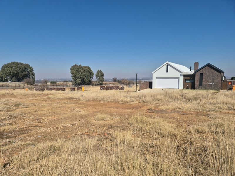 A view of a vacant residential plot featuring dry grass, with a modern brick and white-finished garage structure visible in the background.