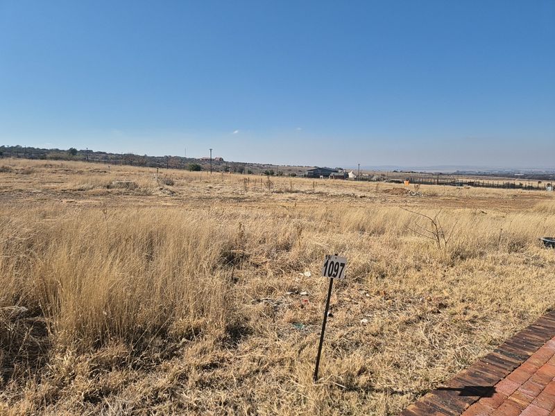 A view of a vacant residential plot featuring dry grass, boundary fencing, and a clear view of the surrounding landscape.