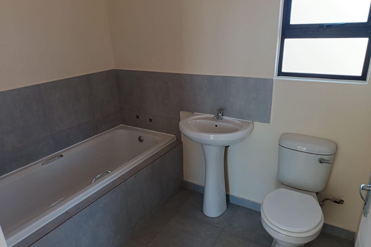 A functional bathroom featuring a bathtub with tiled surround, a pedestal basin, and a standard toilet, finished with neutral wall paint and grey floor tiles.