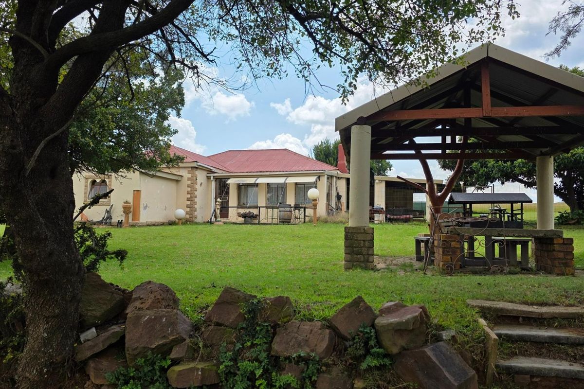 Spacious rear garden featuring a covered outdoor entertainment area with a built-in stone braai and a view of the main farmhouse residence.