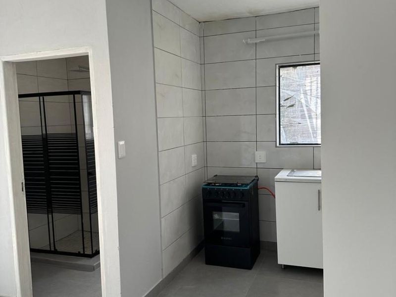 A compact kitchen area featuring tiled walls and flooring, equipped with a freestanding stove and sink cabinet, adjacent to a separate shower room.