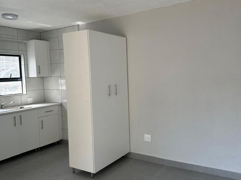 A clean, minimalist kitchen area featuring neutral grey floor tiles, white cabinetry, and a stainless steel sink with a tiled backsplash.