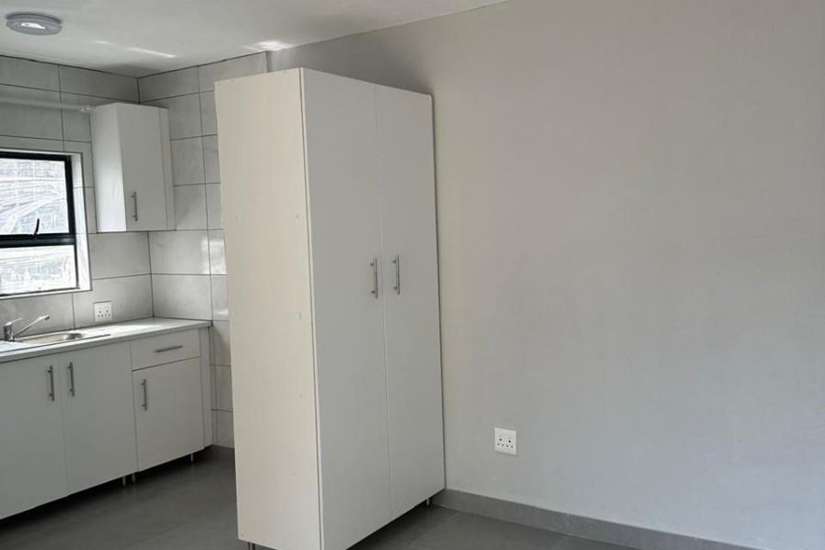 A clean, minimalist kitchen area featuring neutral grey floor tiles, white cabinetry, and a stainless steel sink with a tiled backsplash.