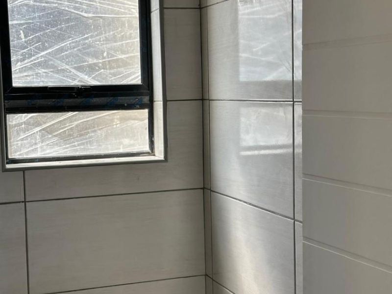 A clean, modern bathroom featuring neutral wall tiles, a white toilet, and a window with an aluminium frame.