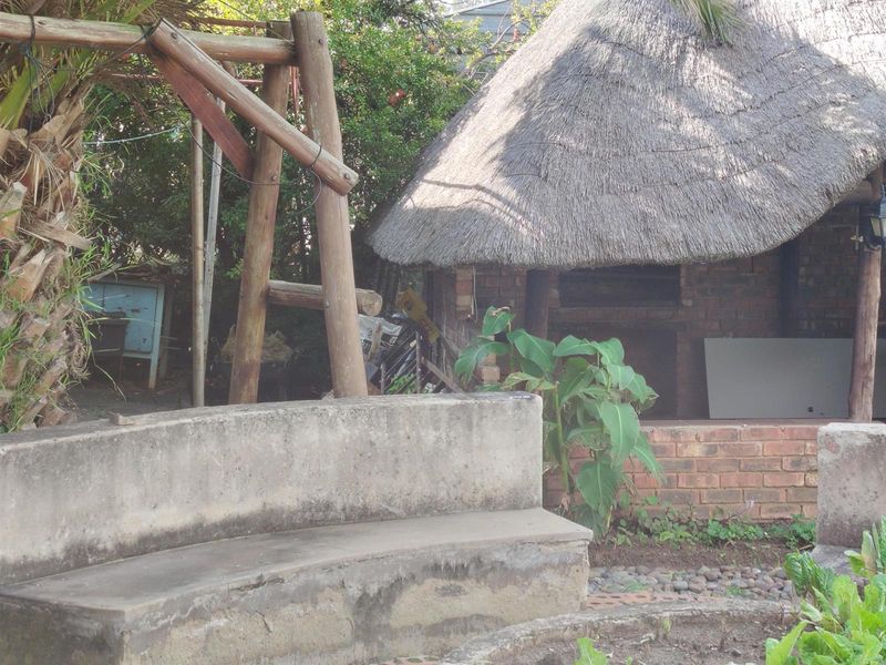 Outdoor entertainment area featuring a thatched lapa with a built-in brick braai and a weathered concrete bench in a garden setting.