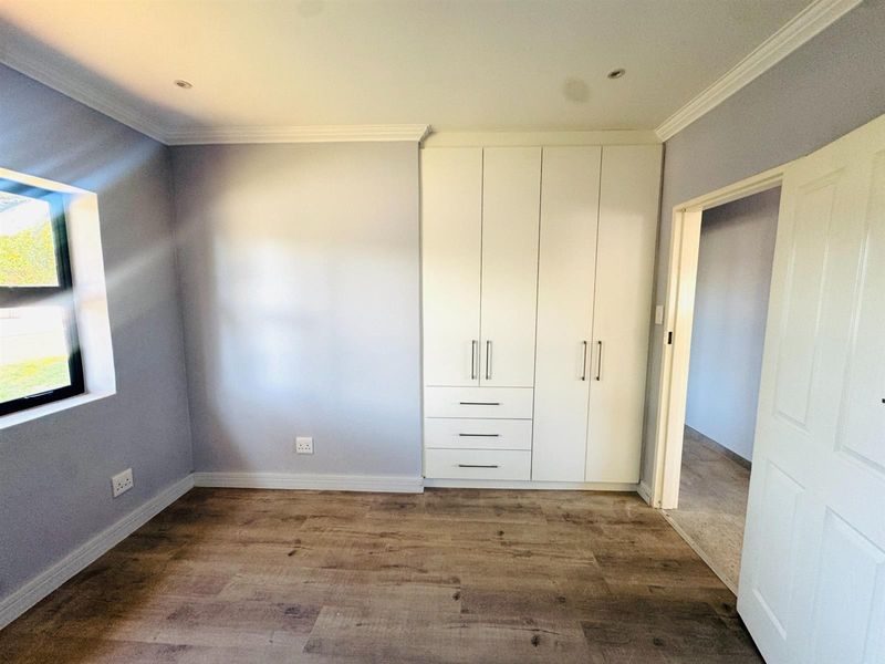 A bright, empty bedroom featuring modern laminate flooring, built-in white cupboards, recessed downlights, and neutral wall finishes.