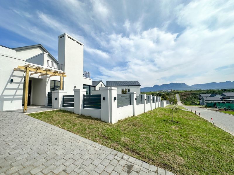 Modern residential exterior featuring a paved driveway, timber pergola, and secure masonry boundary wall with metal fencing, set against a scenic mountain backdrop.