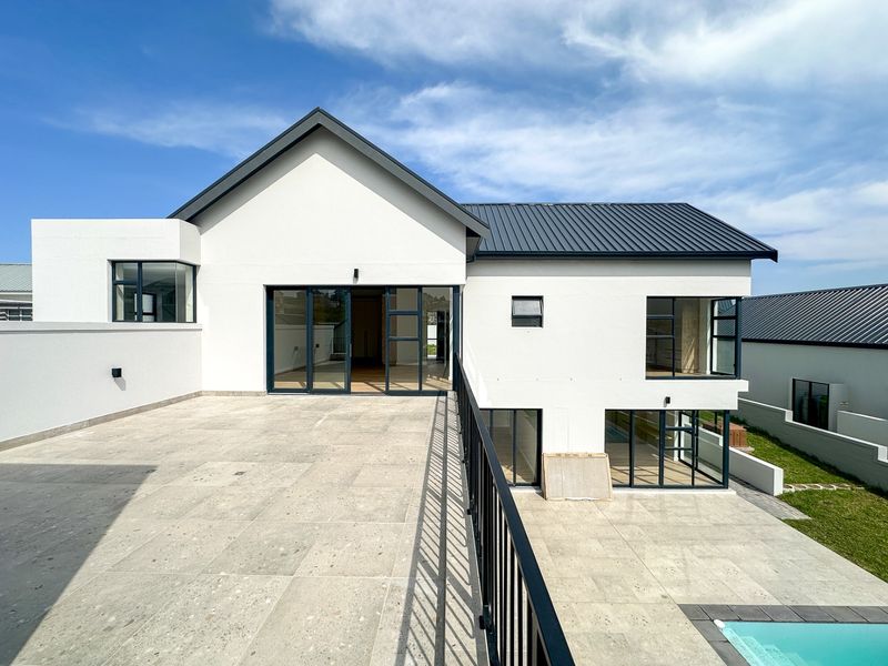 A modern rear elevation featuring a spacious tiled patio, metal roofing, large sliding glass doors, and a swimming pool with a sleek metal balustrade.