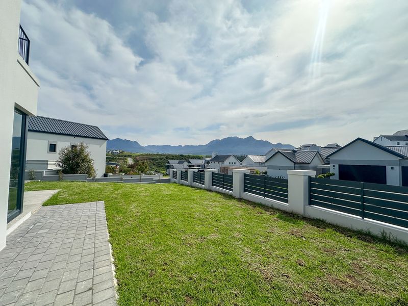 A well-maintained rear garden featuring a paved walkway, manicured lawn, and modern boundary wall with metal fencing, offering scenic mountain views.
