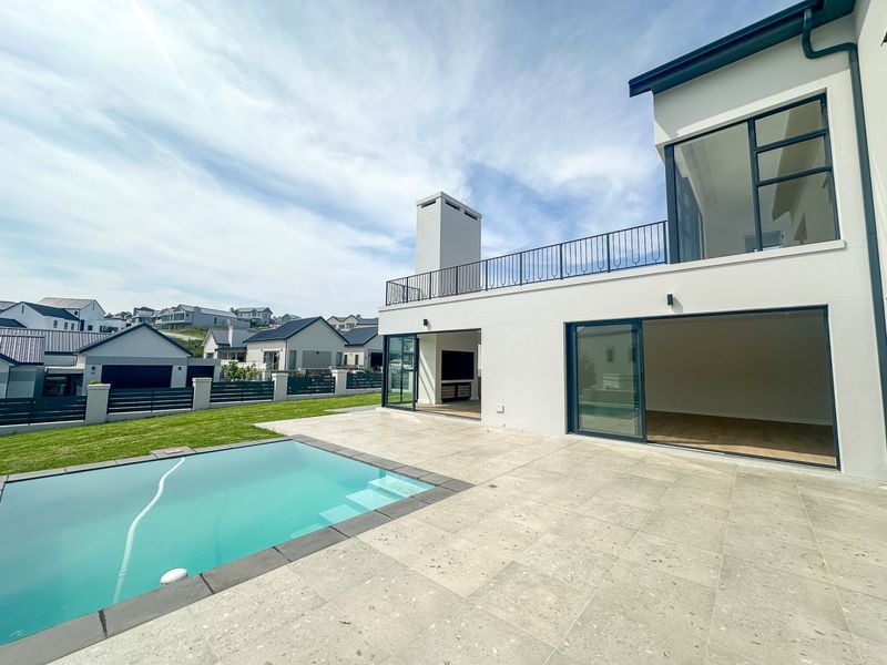 Rear exterior view featuring a modern home with a swimming pool, stone patio, and an upper-level balcony with metal railings.