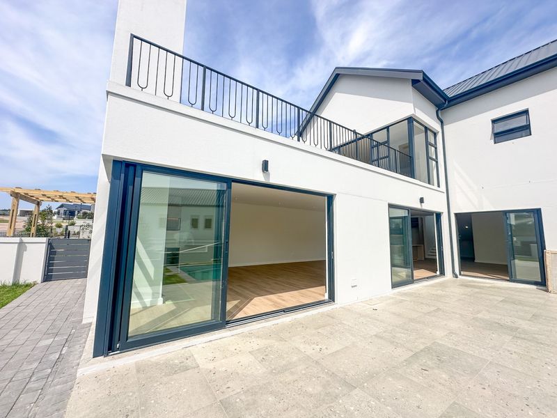 Rear view of a modern home featuring a spacious stone patio, large sliding glass doors, and an upper-level balcony with metal railings.