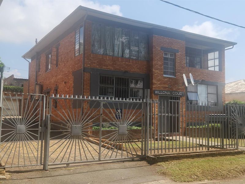 Front elevation of a two-story brick apartment building featuring secure gated access, security bars on windows, and a paved driveway area.