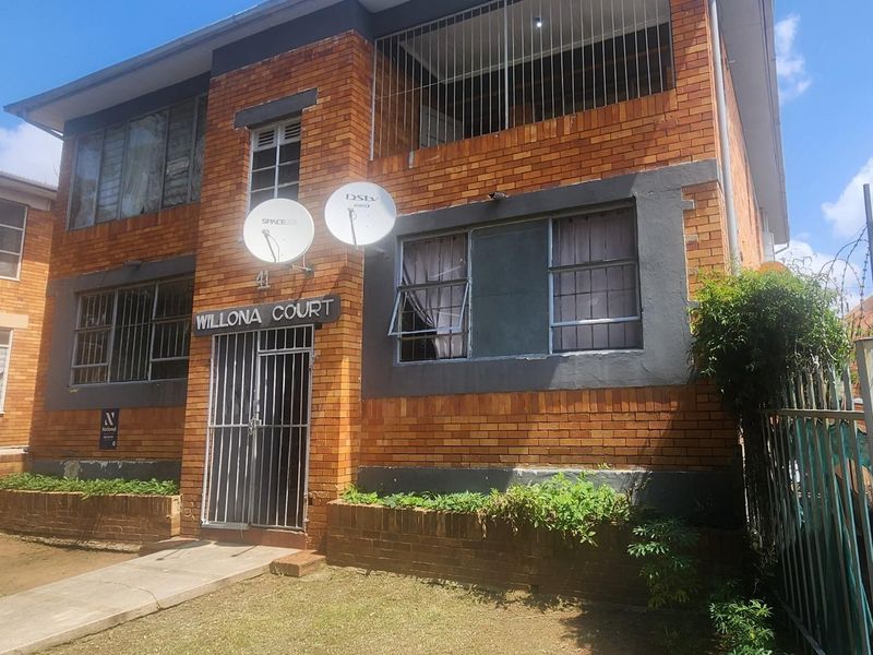 Front elevation of Willona Court, a multi-story face brick apartment building featuring security bars and perimeter fencing.