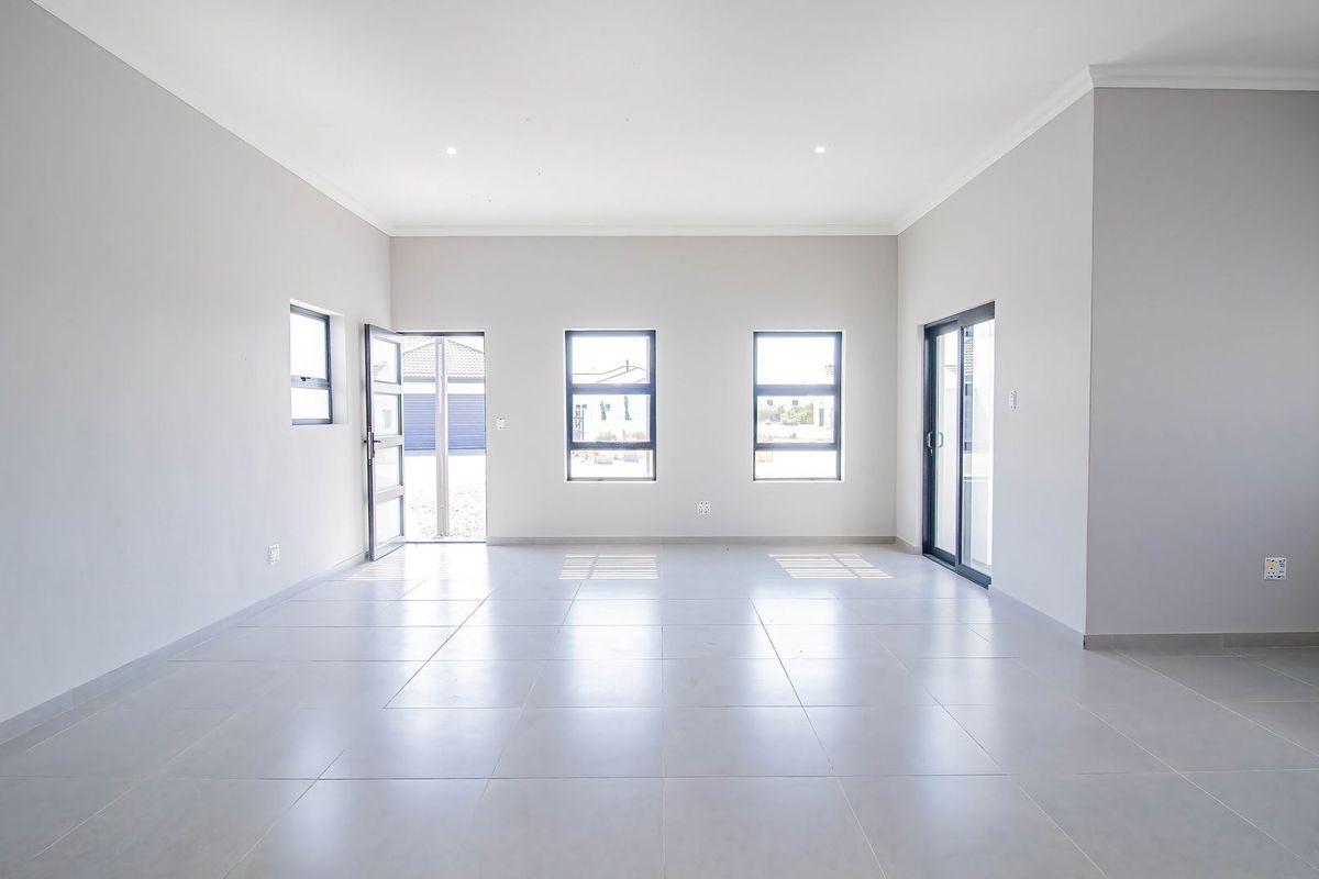 A bright, spacious, and empty living area featuring neutral tiled flooring, modern aluminium window frames, and a sliding glass door leading to the exterior.