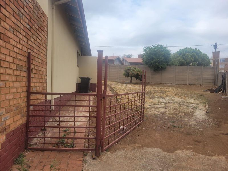 Rear exterior view of the property featuring a brick facade, paved walkway, metal gate, and a large, unlandscaped backyard with boundary walling.