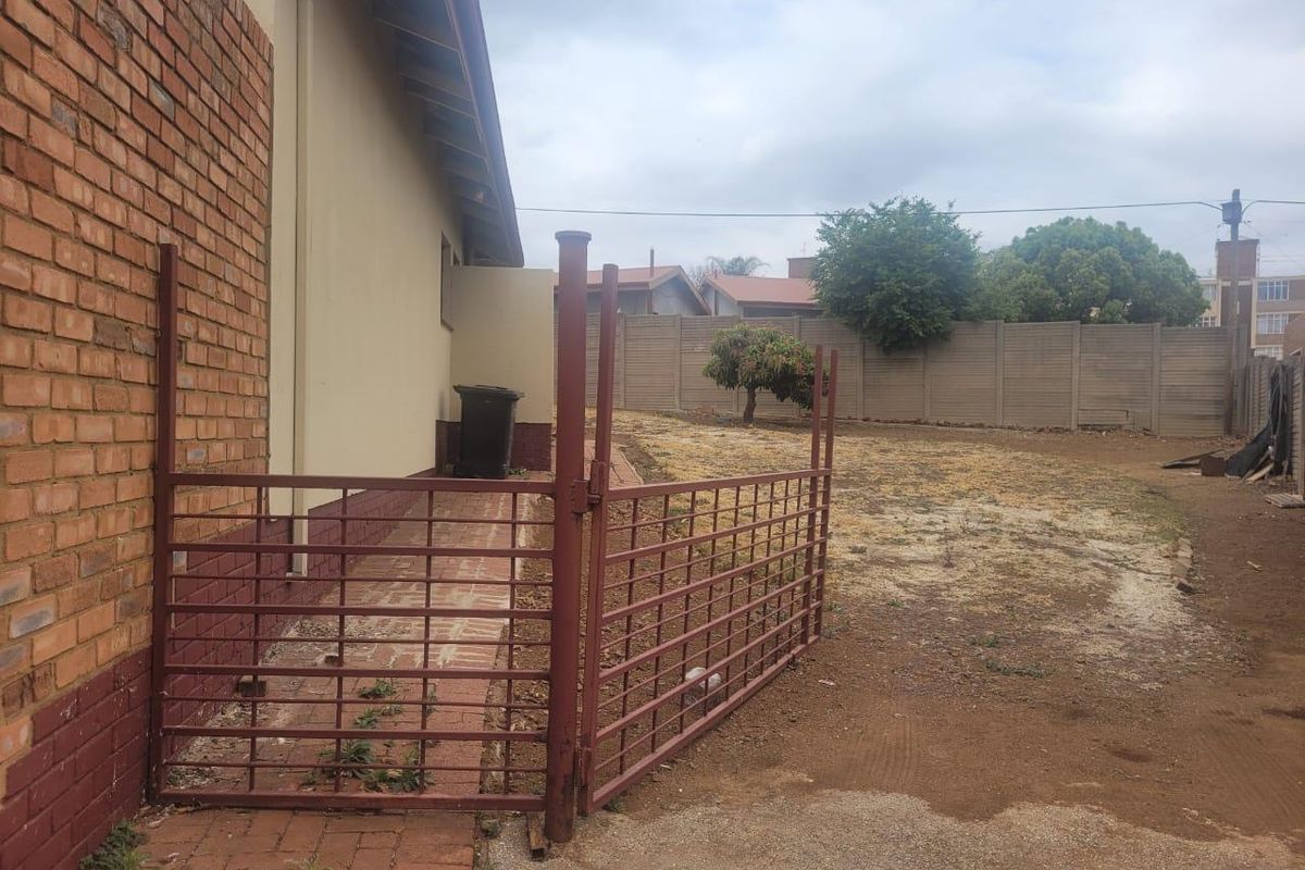 Rear exterior view of the property featuring a brick facade, paved walkway, metal gate, and a large, unlandscaped backyard with boundary walling.