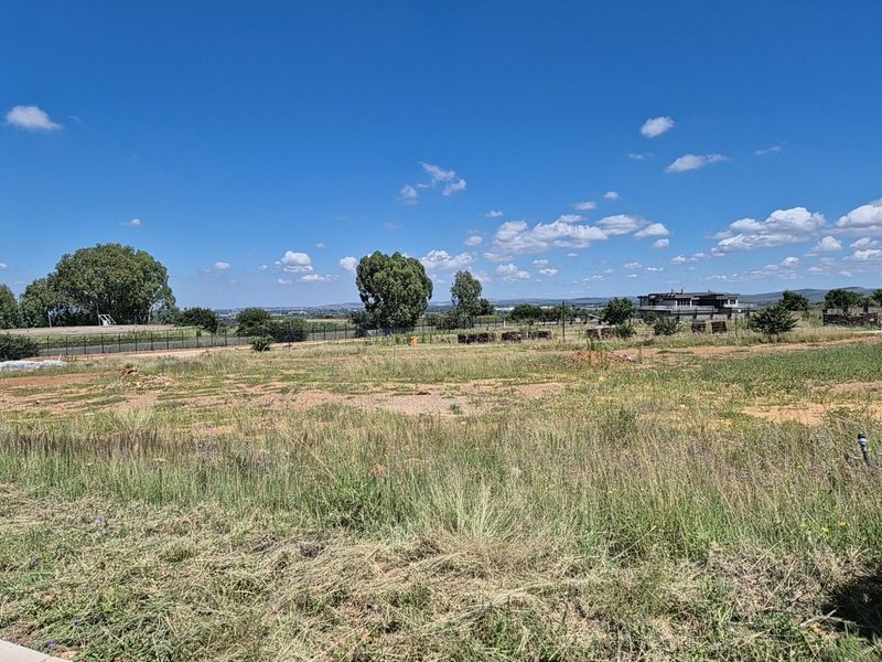 A vacant residential plot of land featuring natural grass cover, perimeter fencing, and distant views of the surrounding neighborhood.