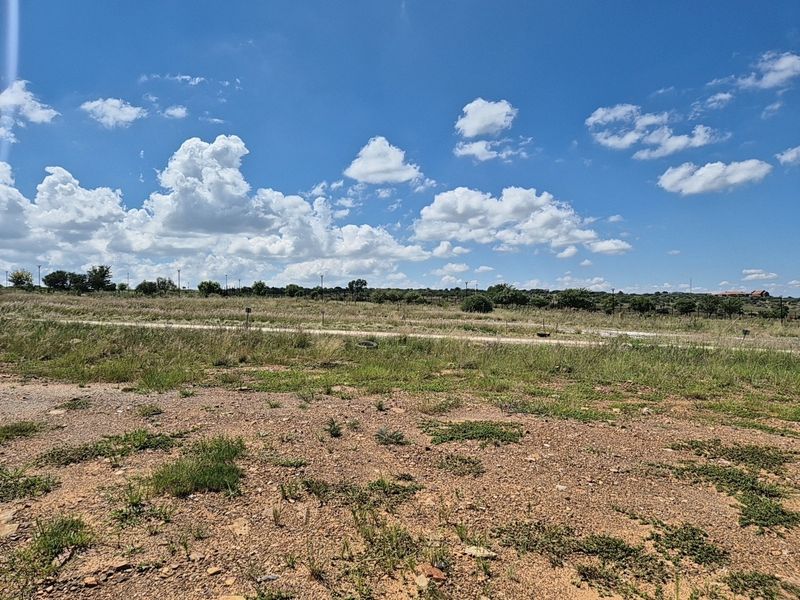 A wide-angle view of a vacant residential plot featuring natural grass and scrub vegetation under a clear blue sky.