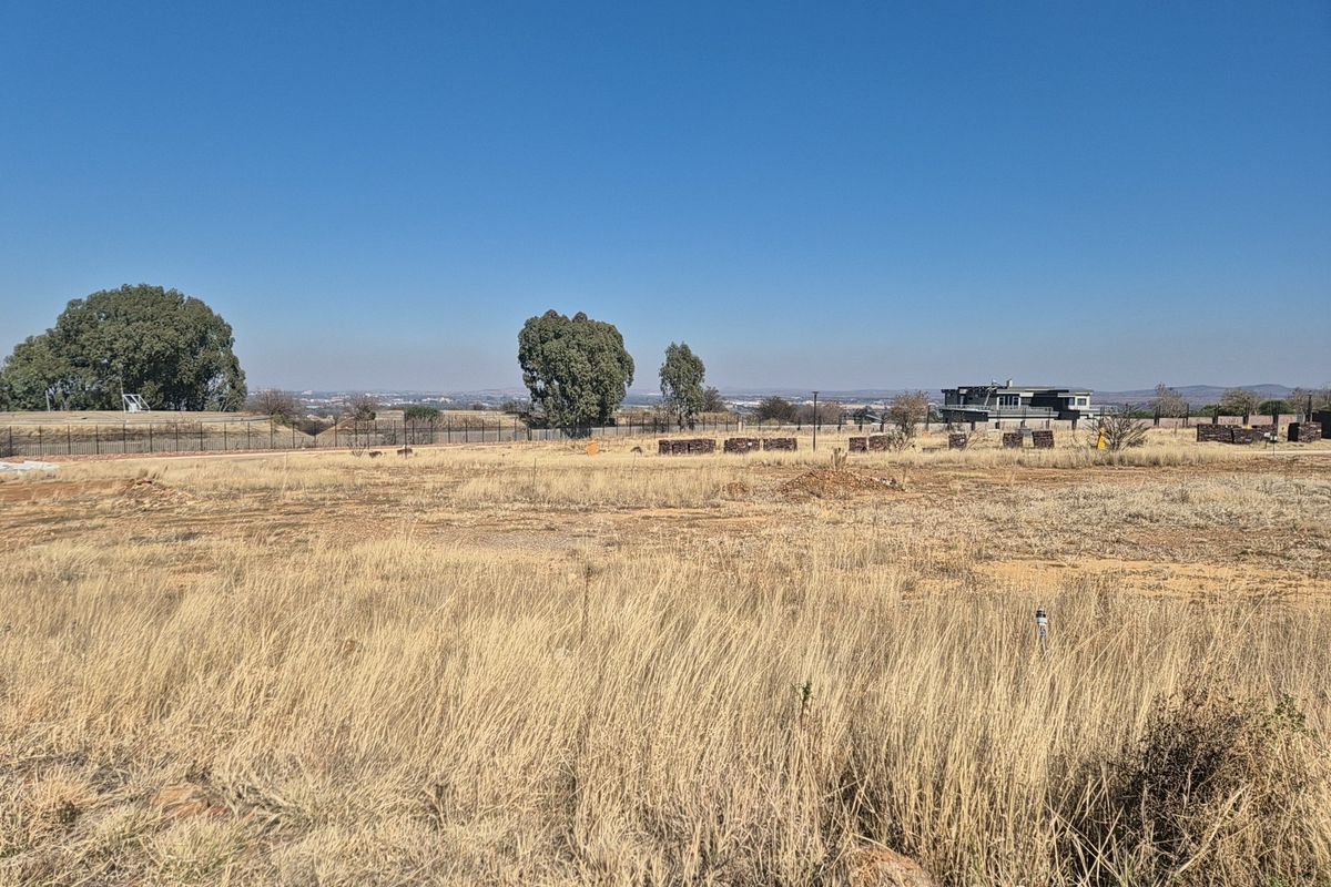 A wide-angle view of a vacant residential plot featuring dry grass, perimeter fencing, and distant neighborhood views under a clear blue sky.