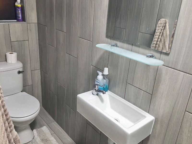 A modern, compact bathroom featuring grey wood-look wall and floor tiles, a wall-mounted basin, and a sleek mirror with a glass shelf.
