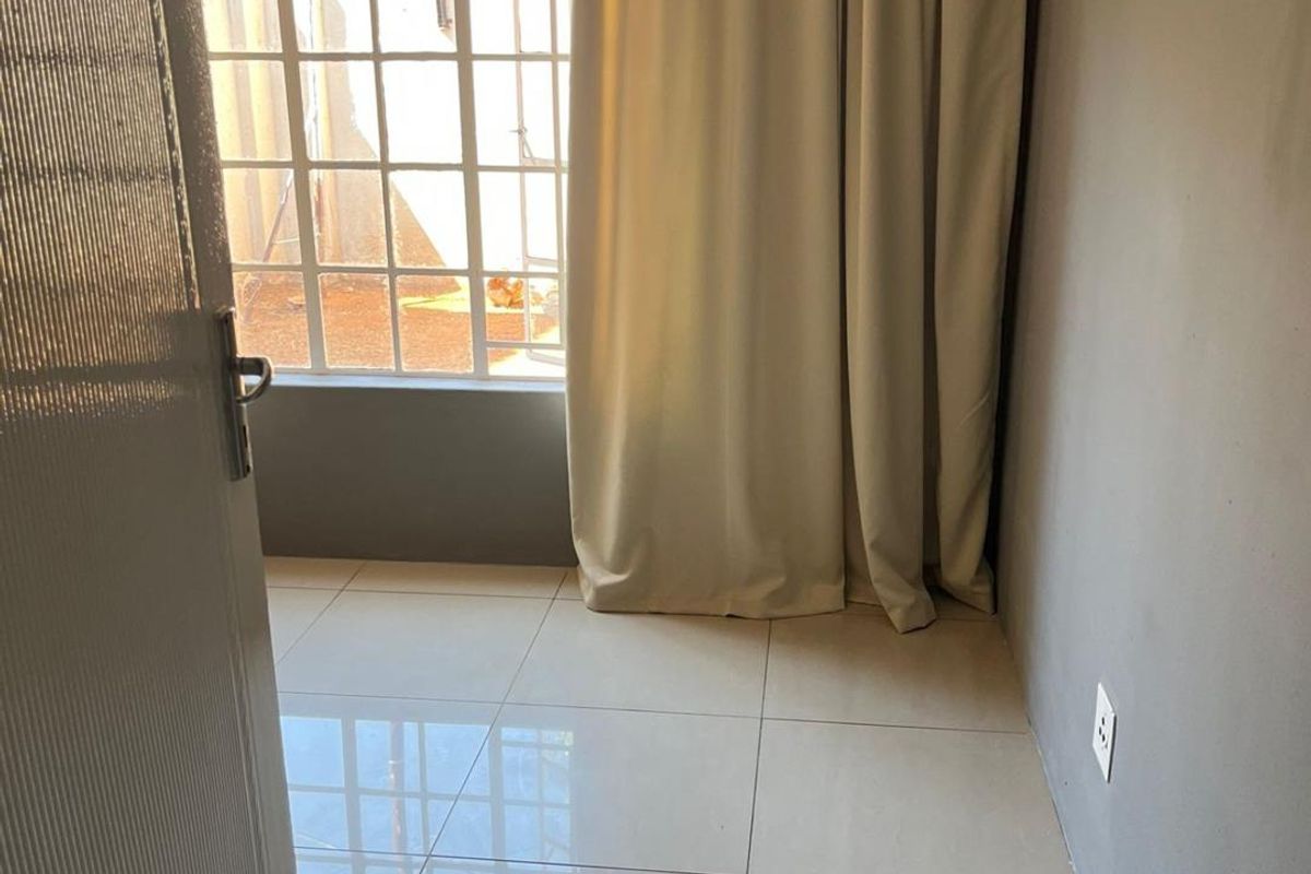 A bright, empty bedroom featuring neutral tiled flooring, grey painted walls, and a large window with curtaining overlooking the exterior area.