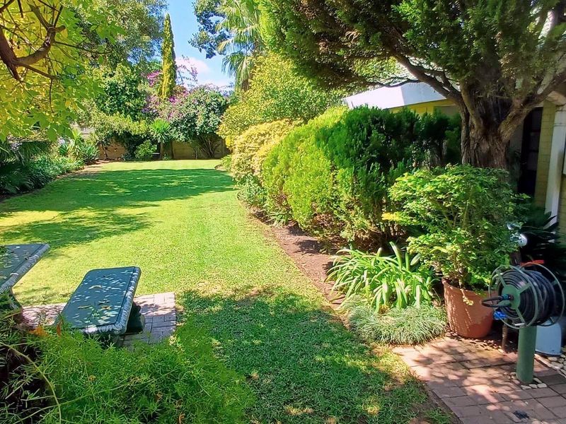 A well-maintained, lush green garden featuring mature trees, manicured hedges, and a paved walkway alongside the property exterior.