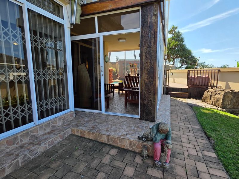 A covered patio area featuring tiled flooring, sliding glass doors with security gates, and access to an adjacent outdoor garden space.