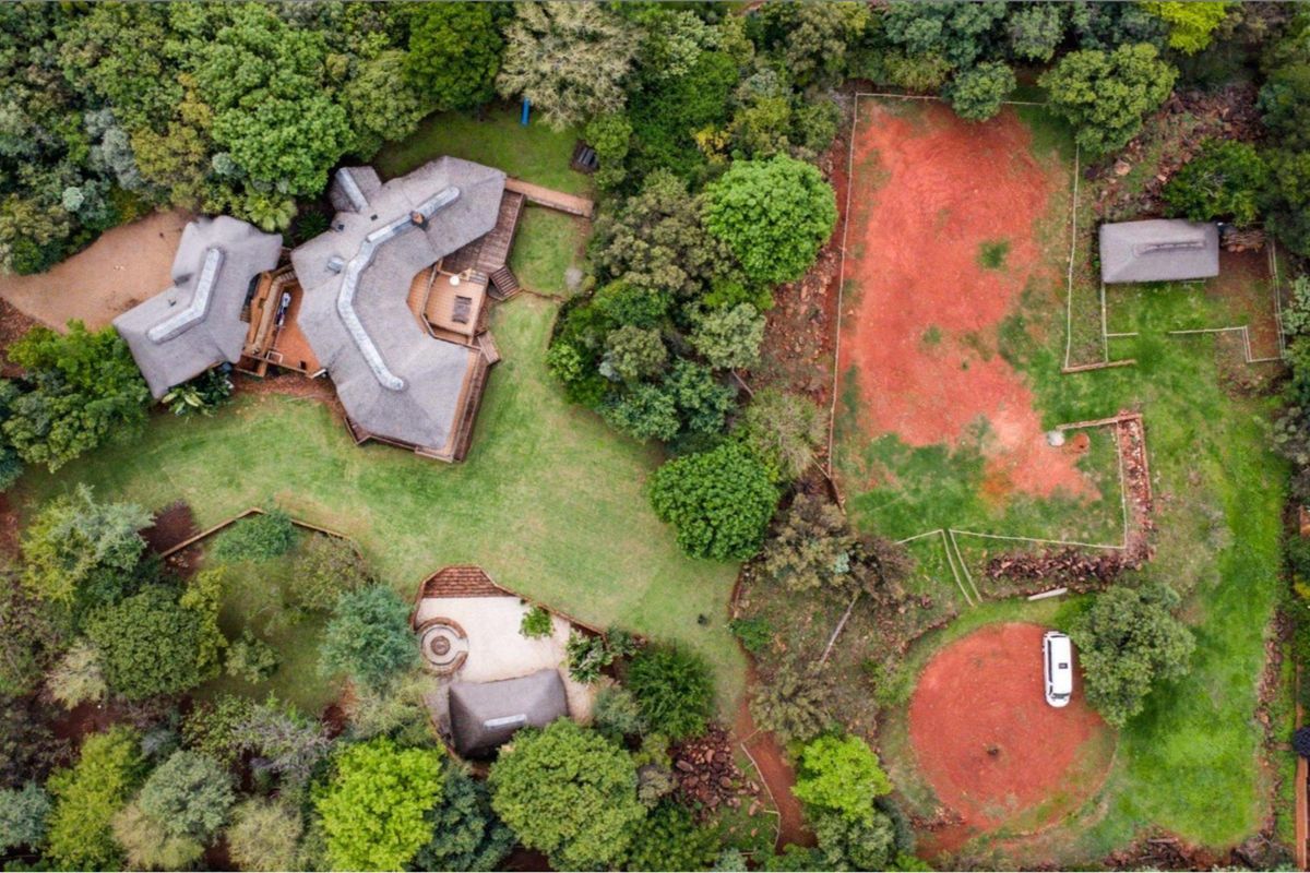 Aerial view of a large property featuring a thatched-roof main house, expansive landscaped gardens, mature trees, and a secondary structure on a spacious plot.