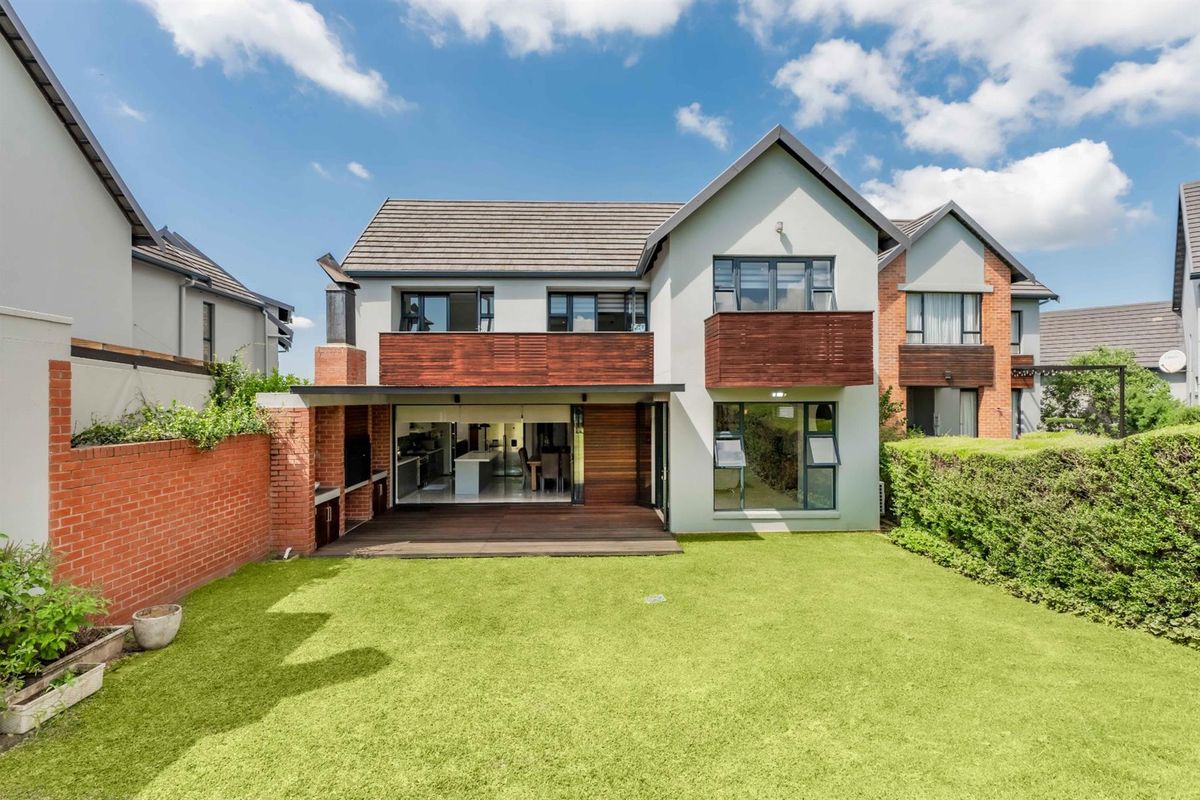 Rear view of a modern townhouse featuring a timber deck, built-in braai area, and a well-maintained private garden with brick boundary walls.