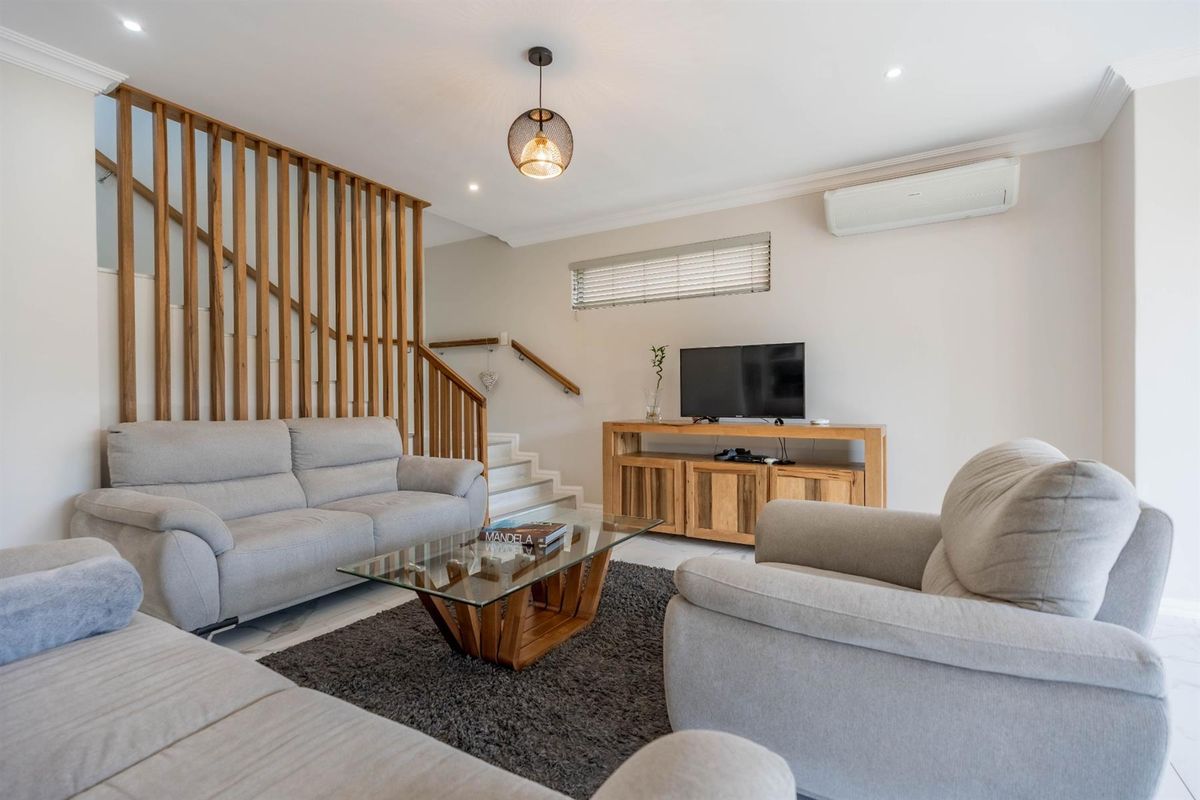 A bright and well-maintained living room featuring a wooden staircase with vertical slat balustrades, recessed lighting, and a wall-mounted air conditioning unit.