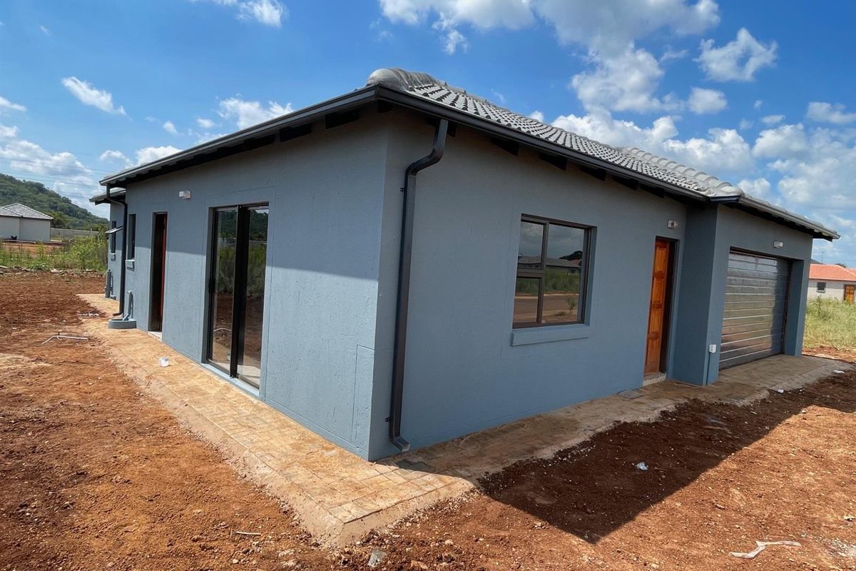 A modern single-story house exterior featuring grey rendered walls, a tiled roof, aluminium window frames, and an integrated garage, set on an unfinished plot.