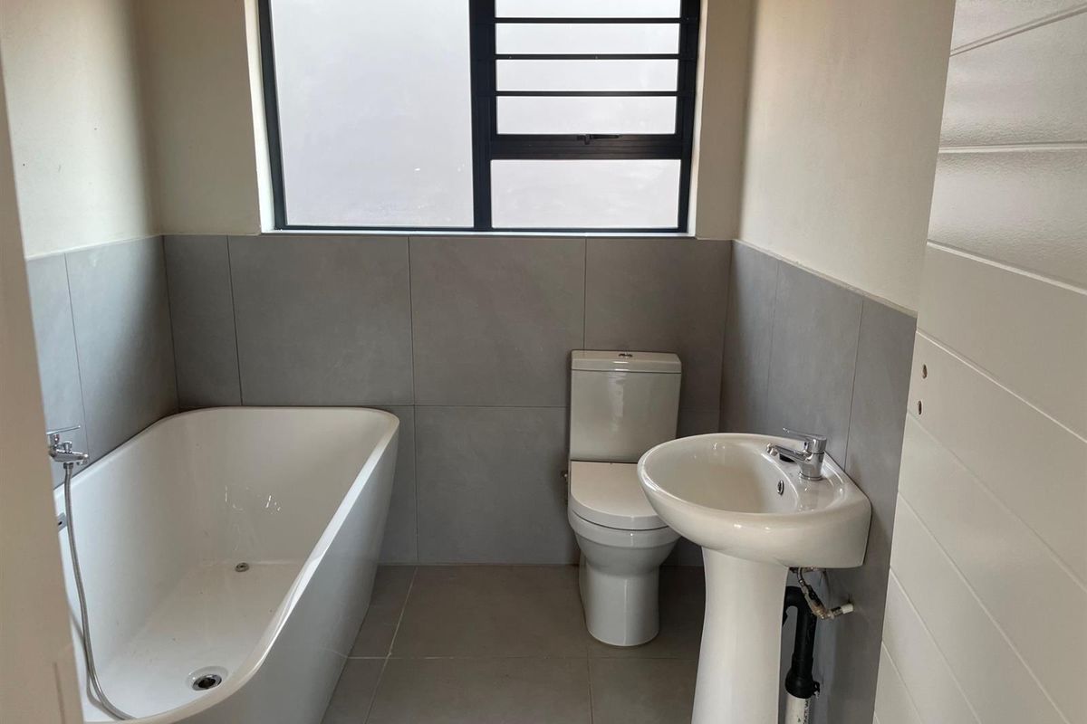 A modern, minimalist bathroom featuring a freestanding bathtub, pedestal basin, and toilet with neutral grey wall and floor tiling.