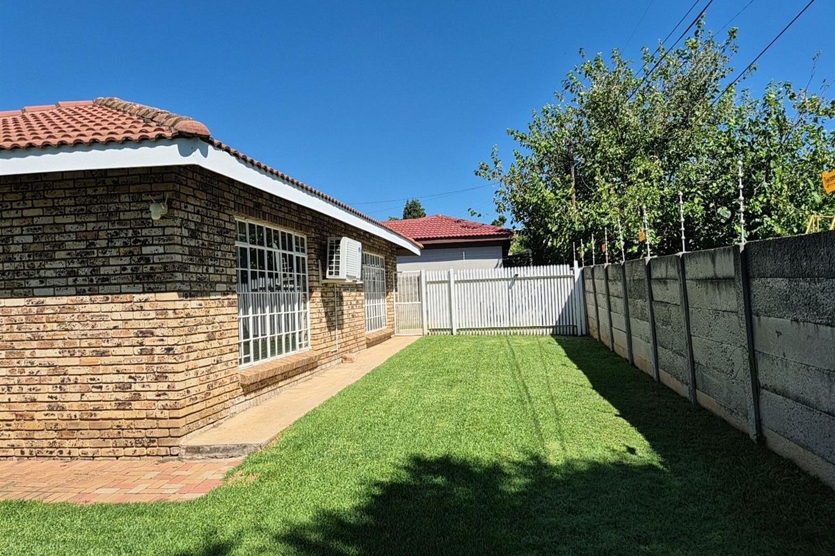 A well-maintained rear garden featuring a brick-faced exterior, paved walkway, manicured lawn, and secure perimeter walling with electric fencing.