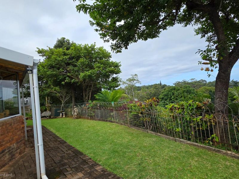 A private rear garden featuring a paved patio area, established lawn, and mature trees, enclosed by security fencing.