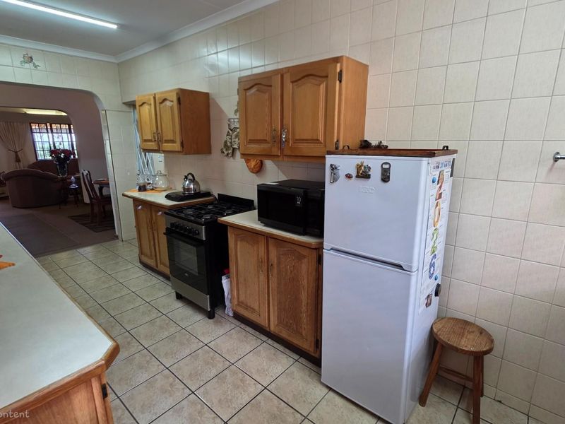 A dated kitchen featuring wooden cabinetry, tiled flooring, and wall-mounted tiling, with a direct view into the adjacent living area.