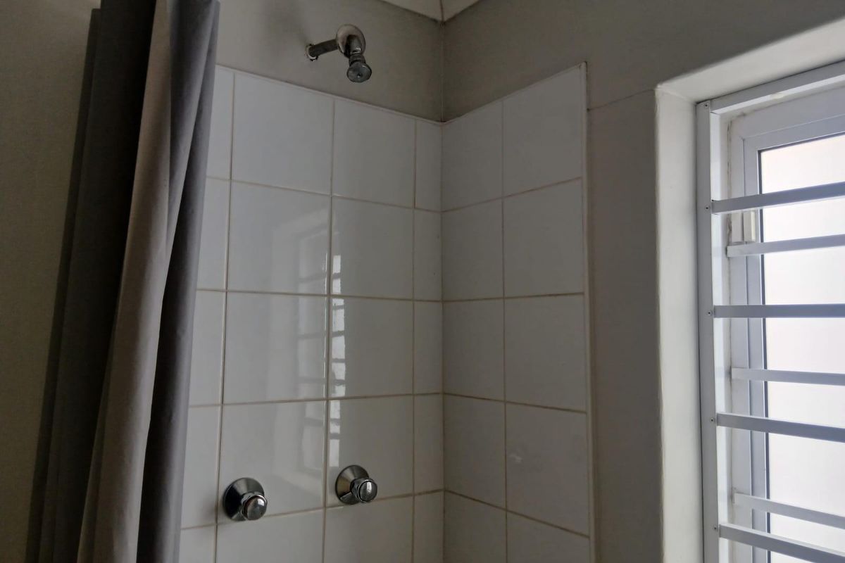 A functional bathroom shower area featuring white ceramic wall tiles, a standard shower head, and a window with security bars.