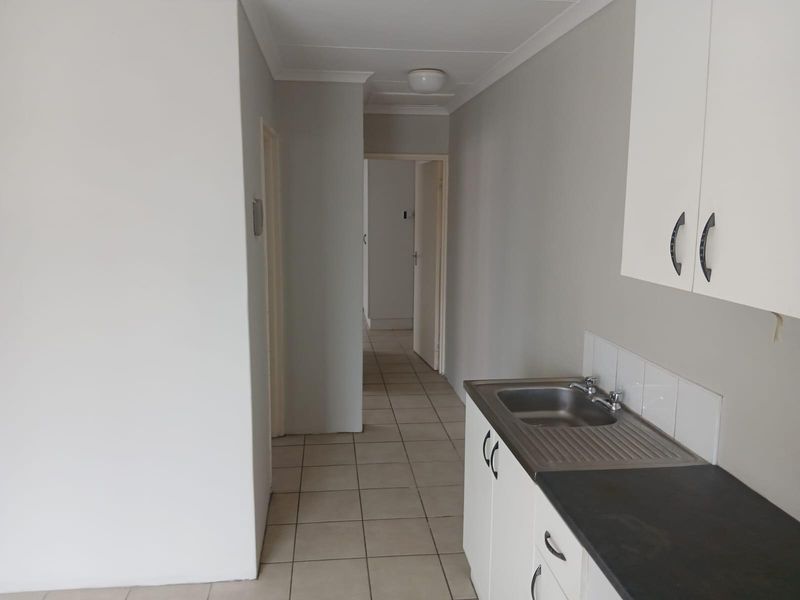 A compact, empty kitchen area featuring built-in cabinetry, a stainless steel sink, and tiled flooring, leading into a hallway.
