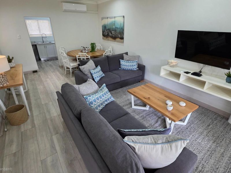 Open-plan living area featuring light wood-look flooring, a wall-mounted media console, and a split-system air conditioning unit, with an adjacent kitchen area.