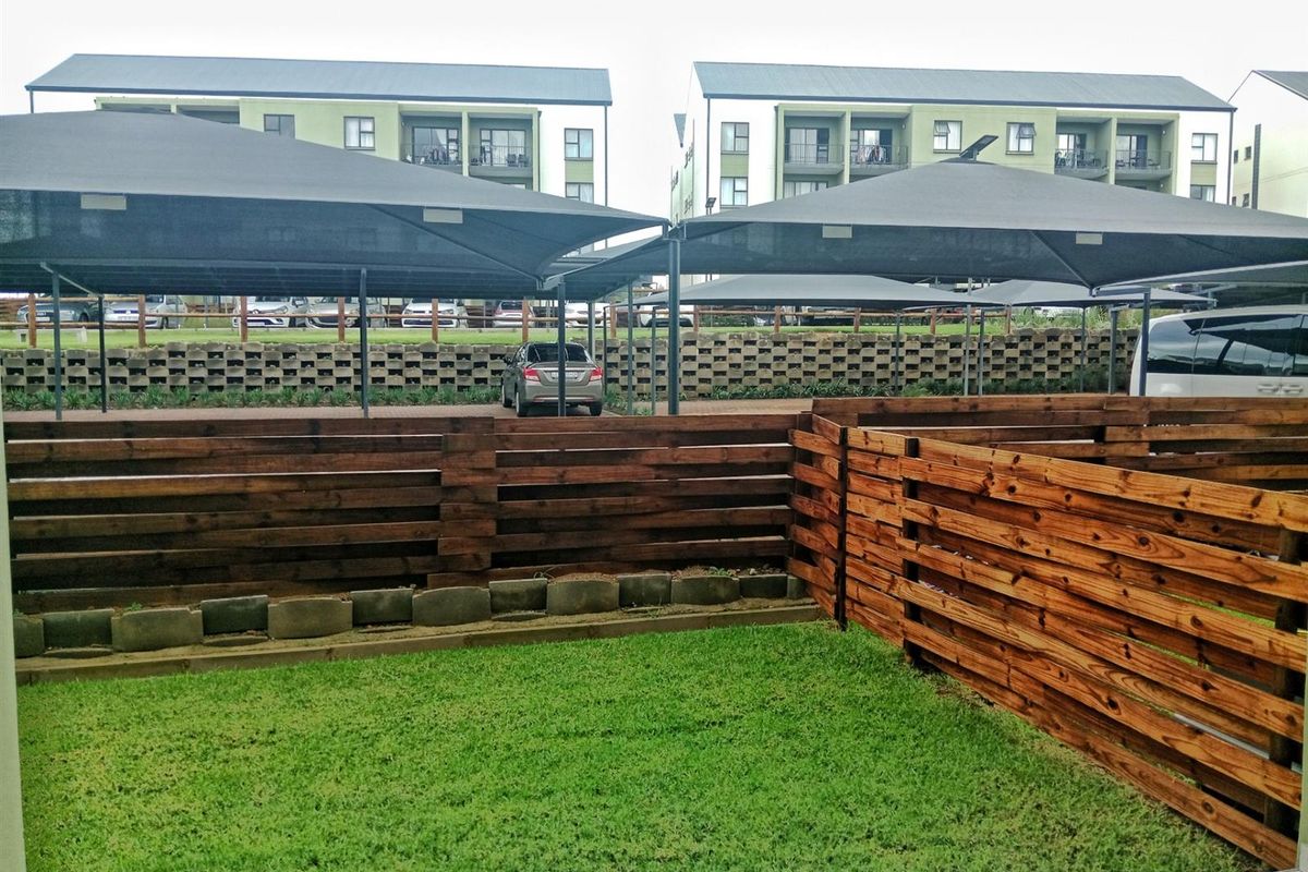 A private rear garden area featuring timber slat fencing, a small lawn, and a view towards the complex parking area with shade cloth carports.