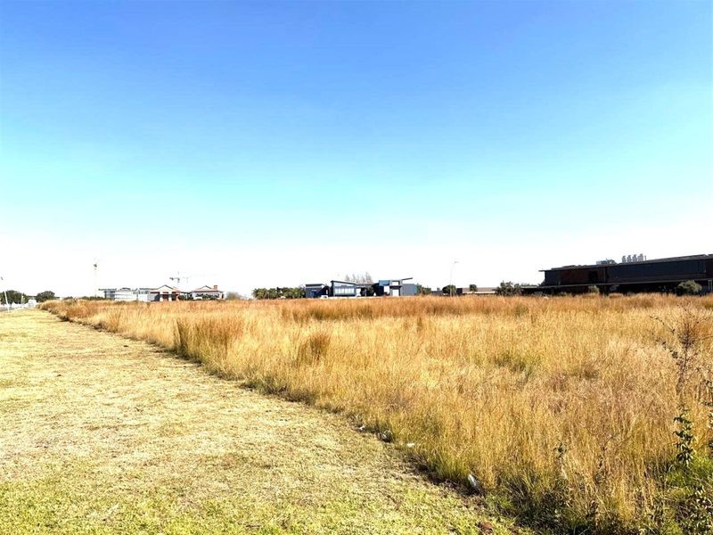 A wide-angle view of a vacant commercial land plot featuring dry grass, with surrounding modern commercial buildings visible in the distance.
