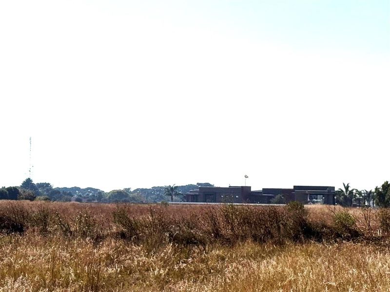 A wide-angle view of a vacant commercial land plot featuring dry grass and distant building structures.