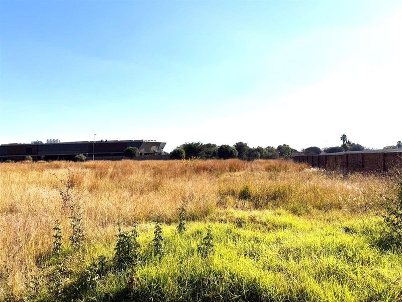 A view of a vacant commercial land plot featuring tall grass, perimeter brick walling, and neighboring commercial structures in the background.