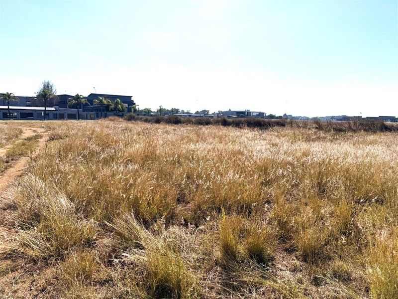 A wide-angle view of a vacant commercial land plot featuring natural grass coverage with established residential developments visible in the background.